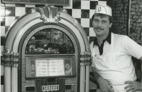 A man in a white uniform and paper hat leans against a classic jukebox in a retro diner with checkered tile walls. He is smiling and has a mustache.
