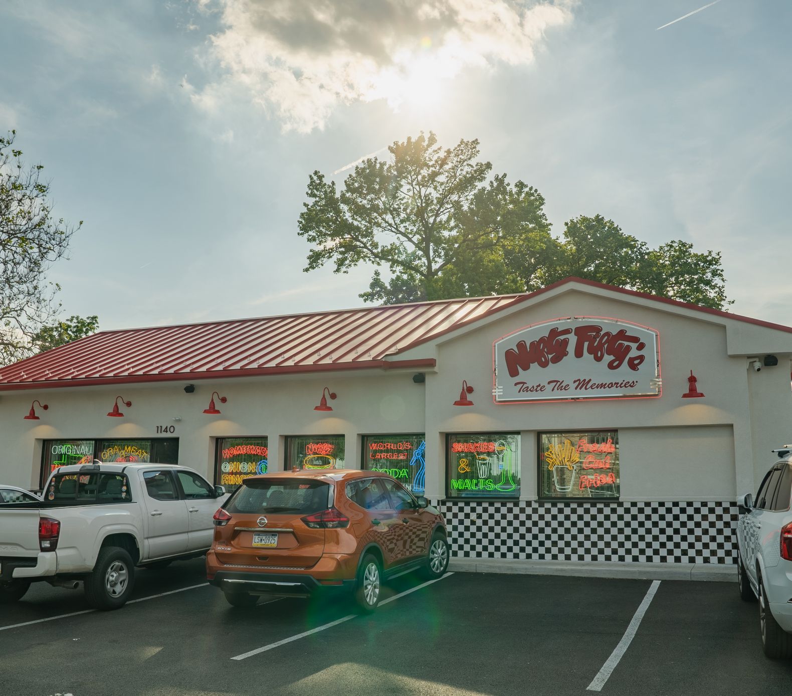 A diner with a red roof and white exterior, featuring a sign that reads Nostalgia: Taste the Memories. Several cars are parked outside, and sunlight streams through the trees behind the building.