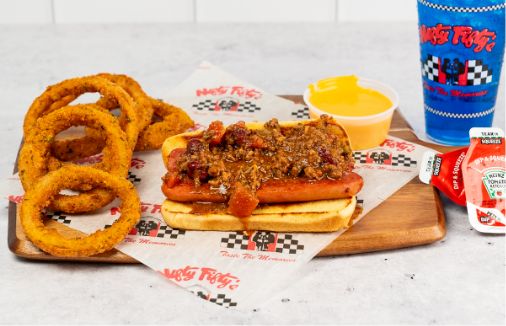 A chili cheese hot dog on a bun with onion rings, a cup of cheese sauce, ketchup and hot sauce packets, and a blue drink cup on a wooden tray. The food is served on paper with a red and black logo.