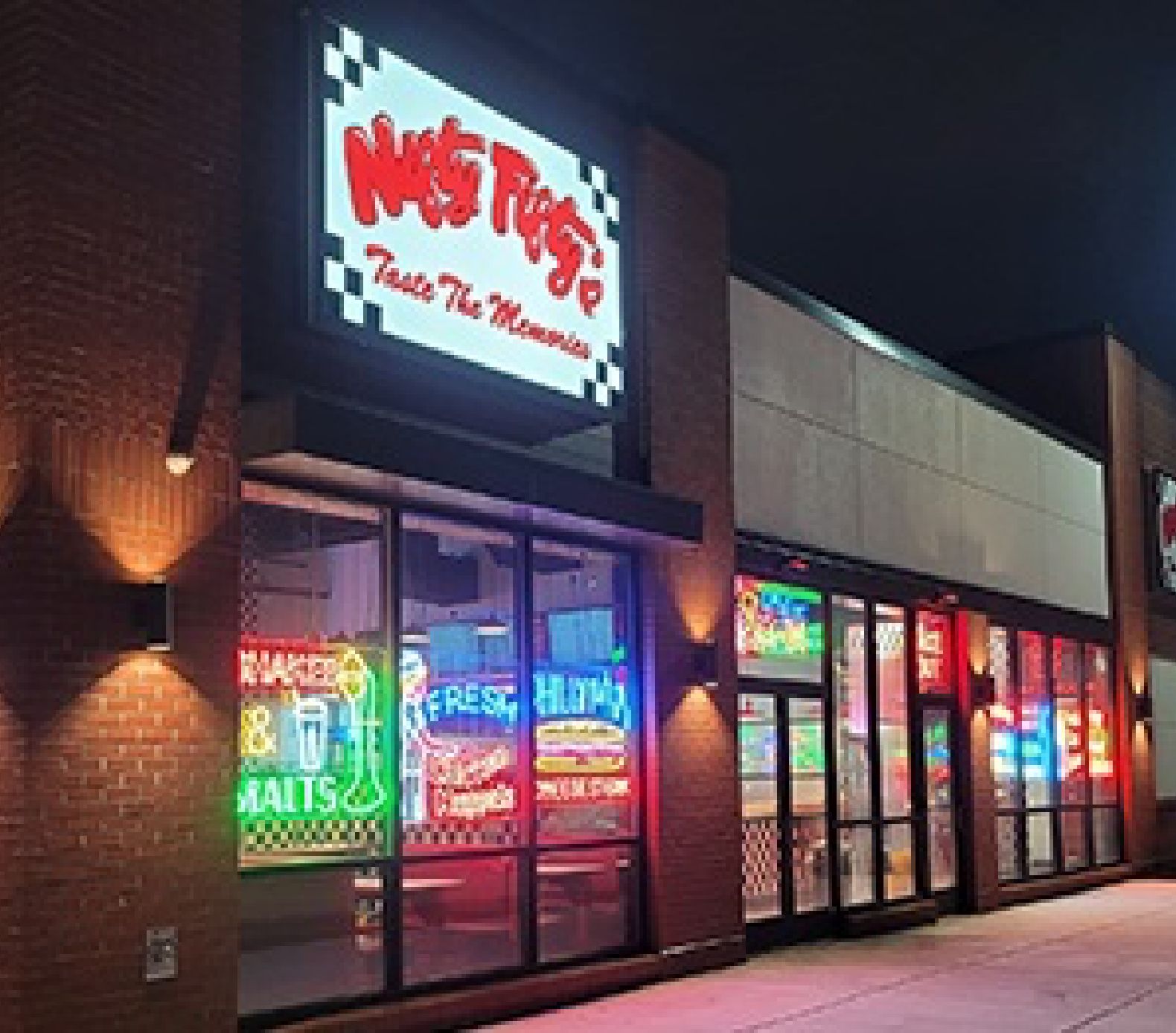 A brightly lit restaurant storefront at night features neon signs in the windows and a large illuminated sign reading “Nifty Fifty’s Taste the Memories” above the entrance.