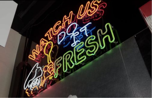 Neon sign on a dark wall reads “WATCH US DO IT FRESH” in red, blue, and green letters, with a white outline of a hand holding a food item on the left side.