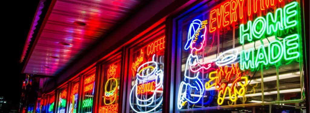 Bright neon signs in a diner window at night, with images of a coffee cup and a person serving food, and text reading EVERYTHING HOME MADE. The colorful lights reflect off the glass and metal awning.