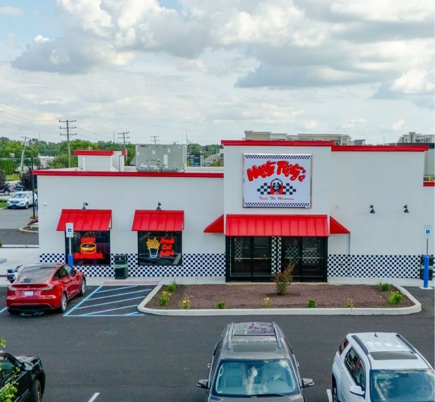 A diner-style restaurant with a red and white exterior, checkered trim, and a sign reading Moe’s Fries. Several cars are parked in front, and the sky is partly cloudy.