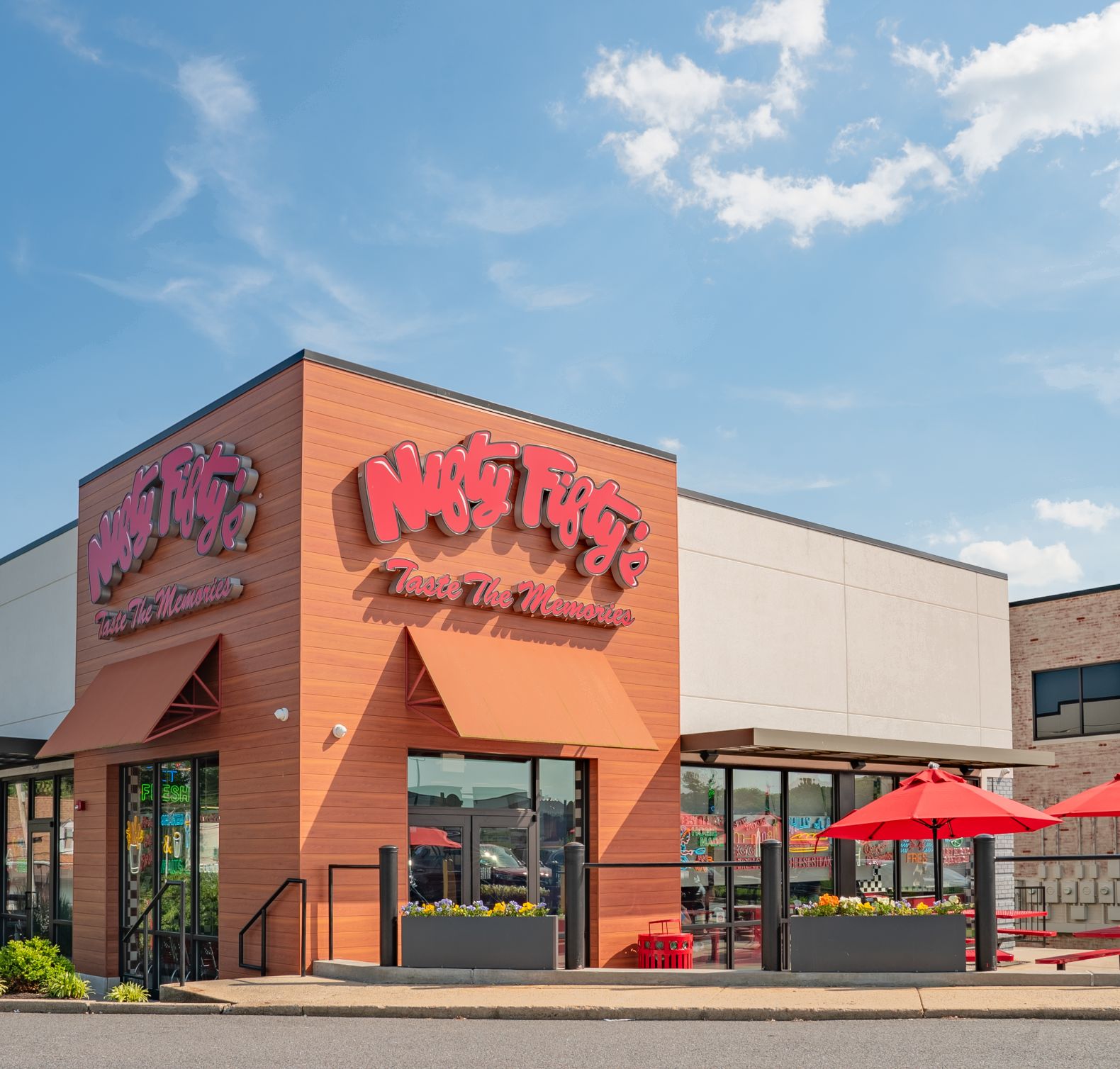 An exterior view of Nifty Fifty's with colorful signage, large windows, flower boxes, and red patio umbrellas on a sunny day with blue sky and clouds.