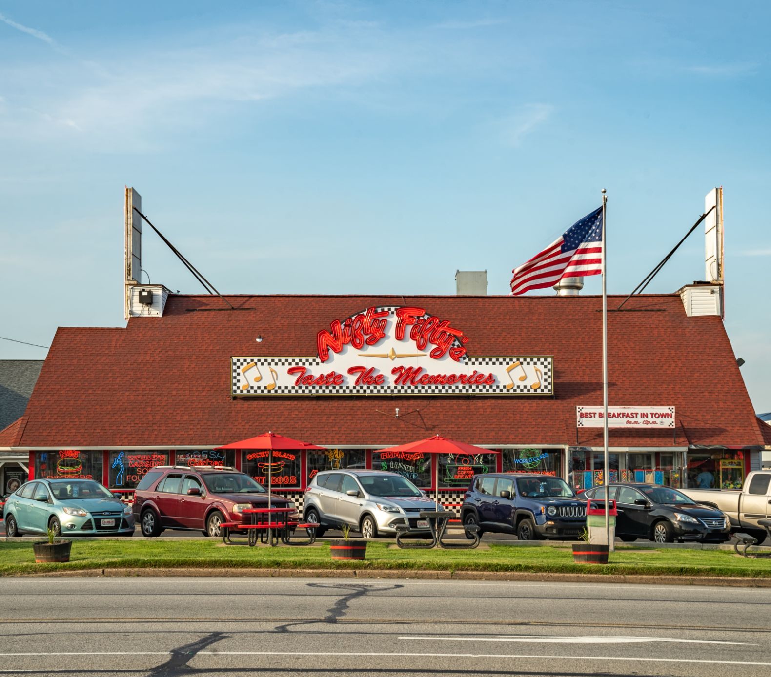 A roadside restaurant with a red roof and large sign reading Mos Place Taste the Memories, an American flag, parked cars, and red patio umbrellas in front.