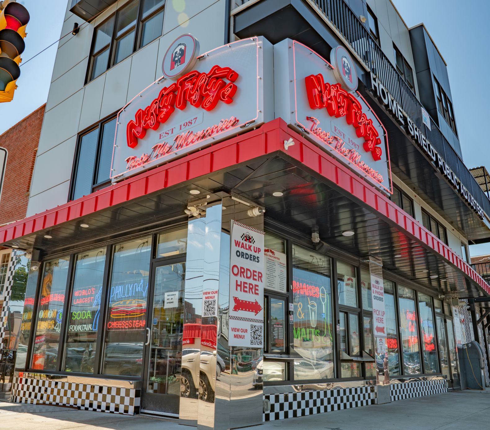 A retro-style diner with checkered tiles and bold red neon signs reading Nifty Fifty's on the corner of a modern building; large windows display bright menu signs inside.